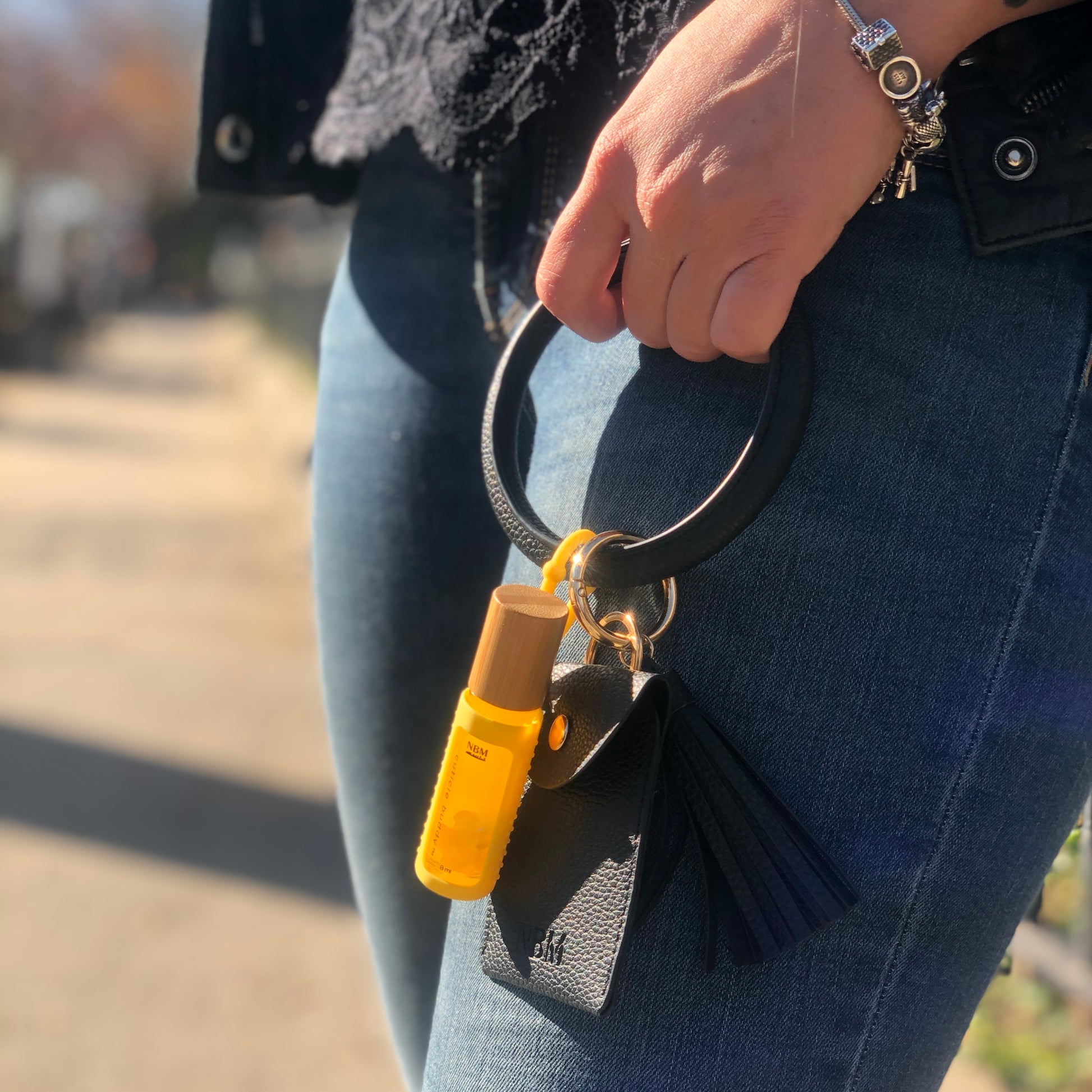 a person wearing a black lace top and dark blue jeans holding a black leather bangle wallet with a bottle of cuticle oil in a yellow silicone holder hanging off the bangle