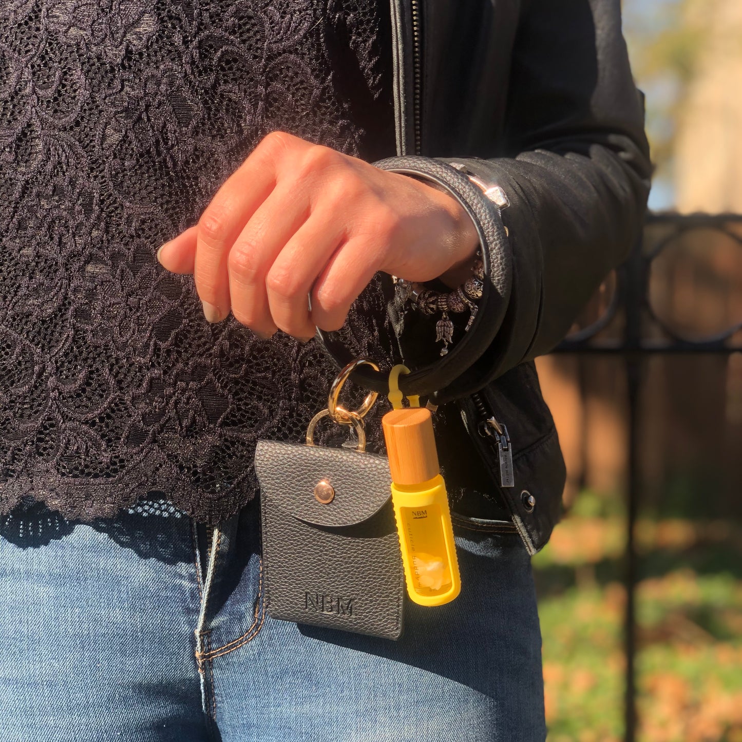 a person wearing a black lace top and dark blue jeans with a black leather bangle wallet hanging off their wrist with a cuticle oil in a yellow silicone holder hanging off the bangle