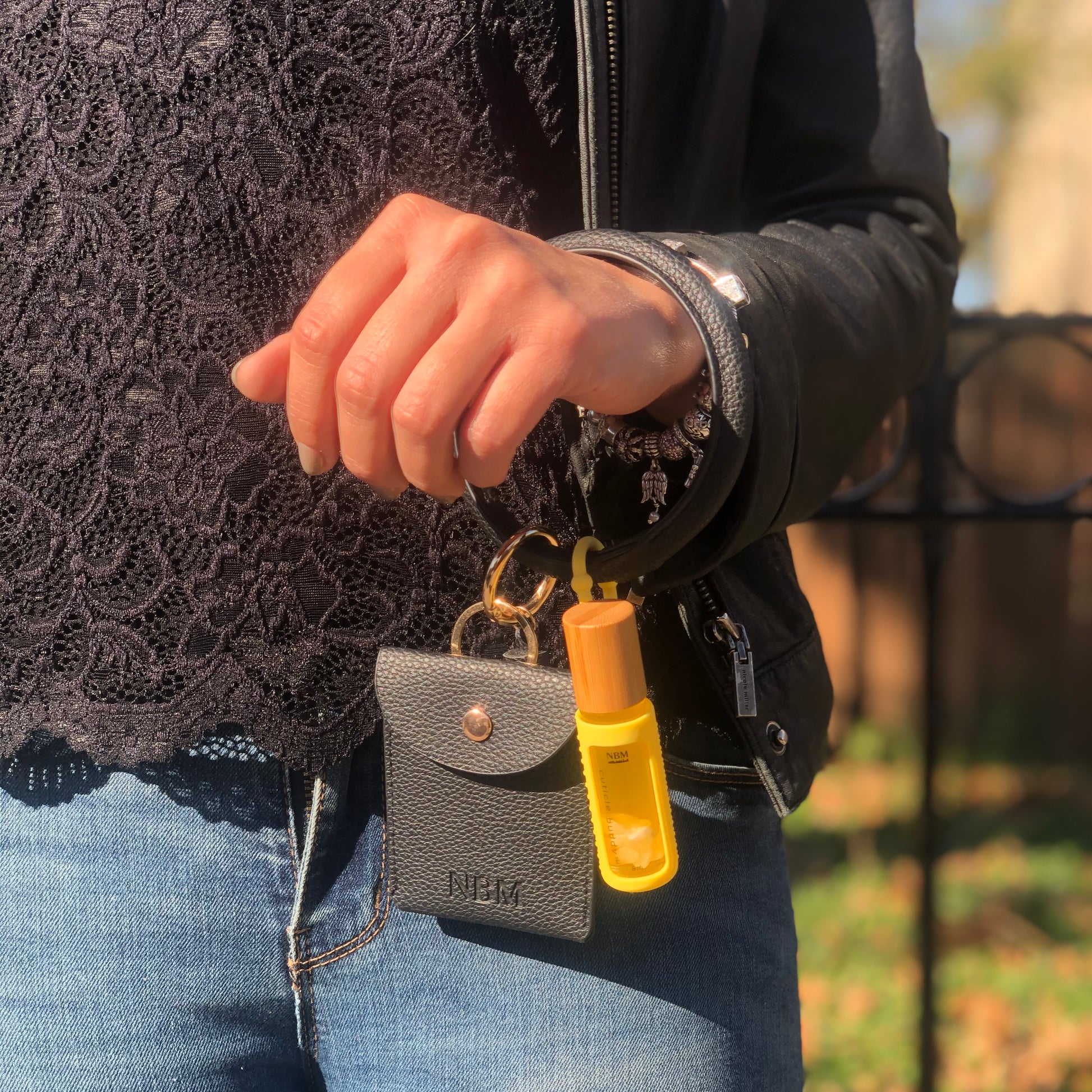 a person wearing a black lace top and dark blue jeans with a black leather bangle wallet hanging off their wrist with a cuticle oil in a yellow silicone holder hanging off the bangle