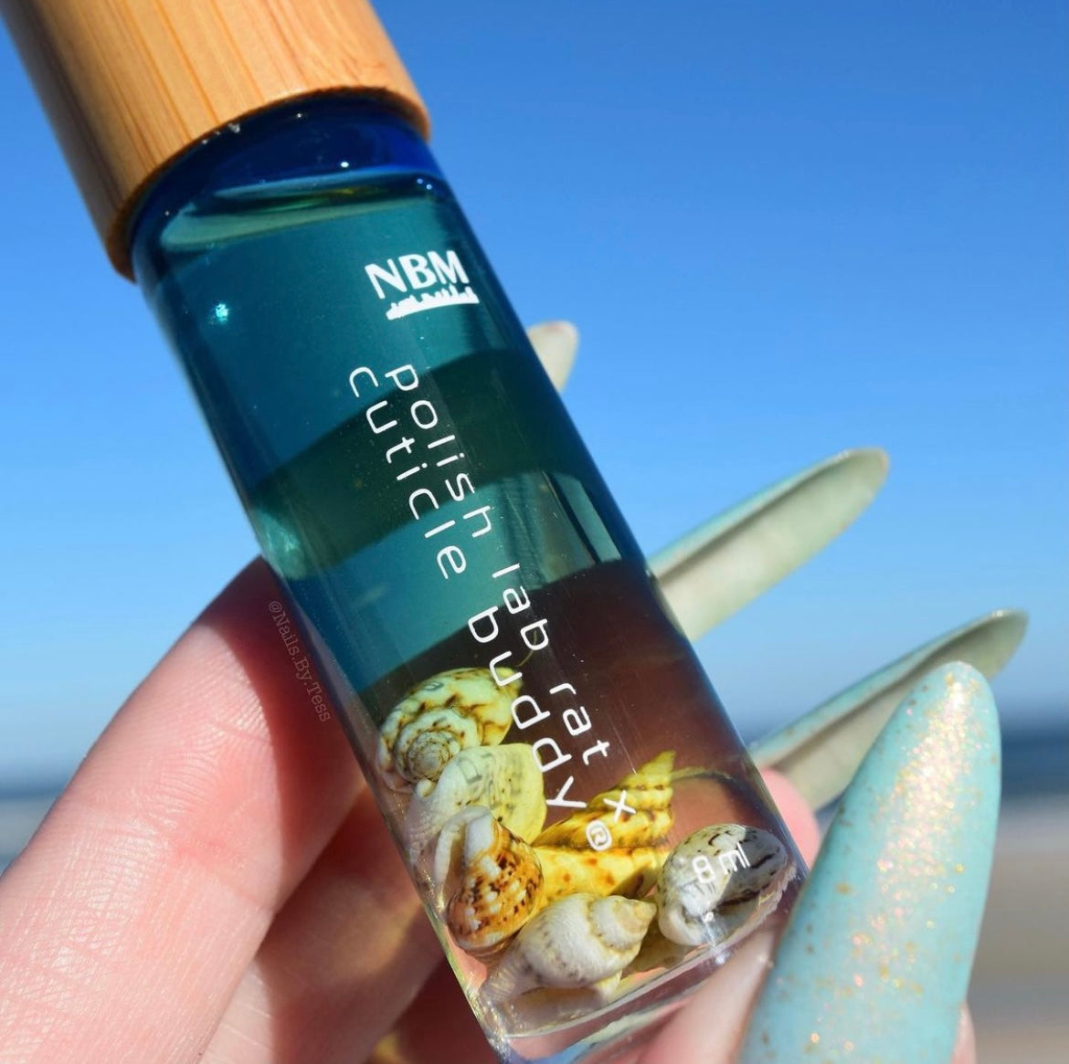 close up shot of a blue tinted bottle of oil being held in a hand with long blue nails. inside the bottle are faux light colored seashells. on a blue sky background.