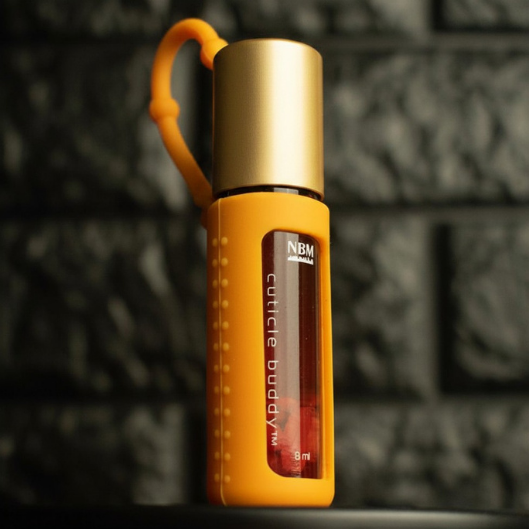 orange rollerball bottle in an orange silicone cover with gold aluminum cap labeled cuticle buddy on a dark textured background