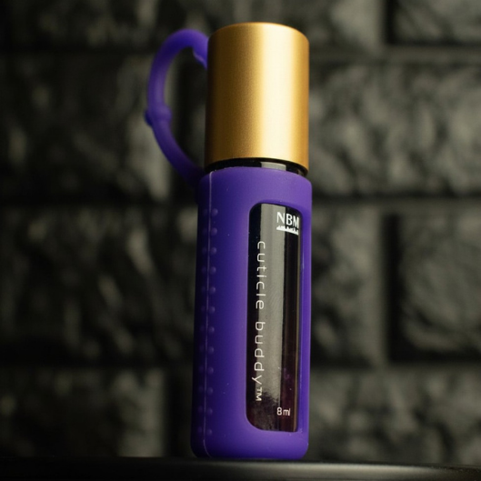 Purple rollerball bottle in a purple silicone cover with gold aluminum cap labeled cuticle buddy on a dark textured background