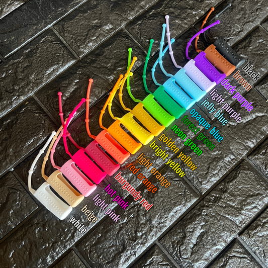 assortment of different colored silicone holders on a black brick background, labeled from left to right: white, beige, light pink, hot pink, burgundy red, red orange, light orange, bright yellow, golden yellow, neon green, forest green, opaque blue, jelly blue, light purple, dark purple, brown, black