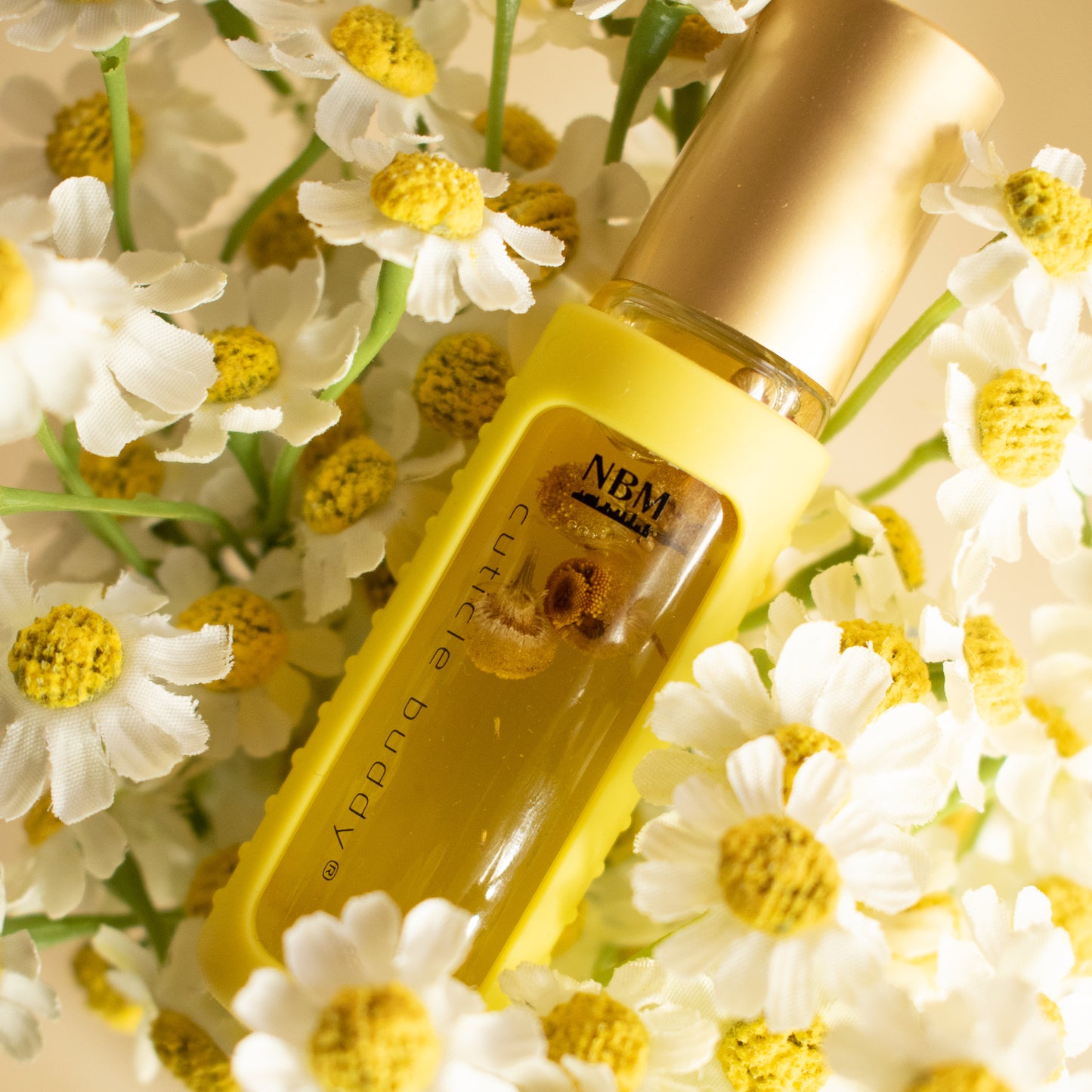 a bottle of cuticle oil with a gold cap in a yellow silicone holder and chamomile buds inside in a bed of faux chamomile flowers