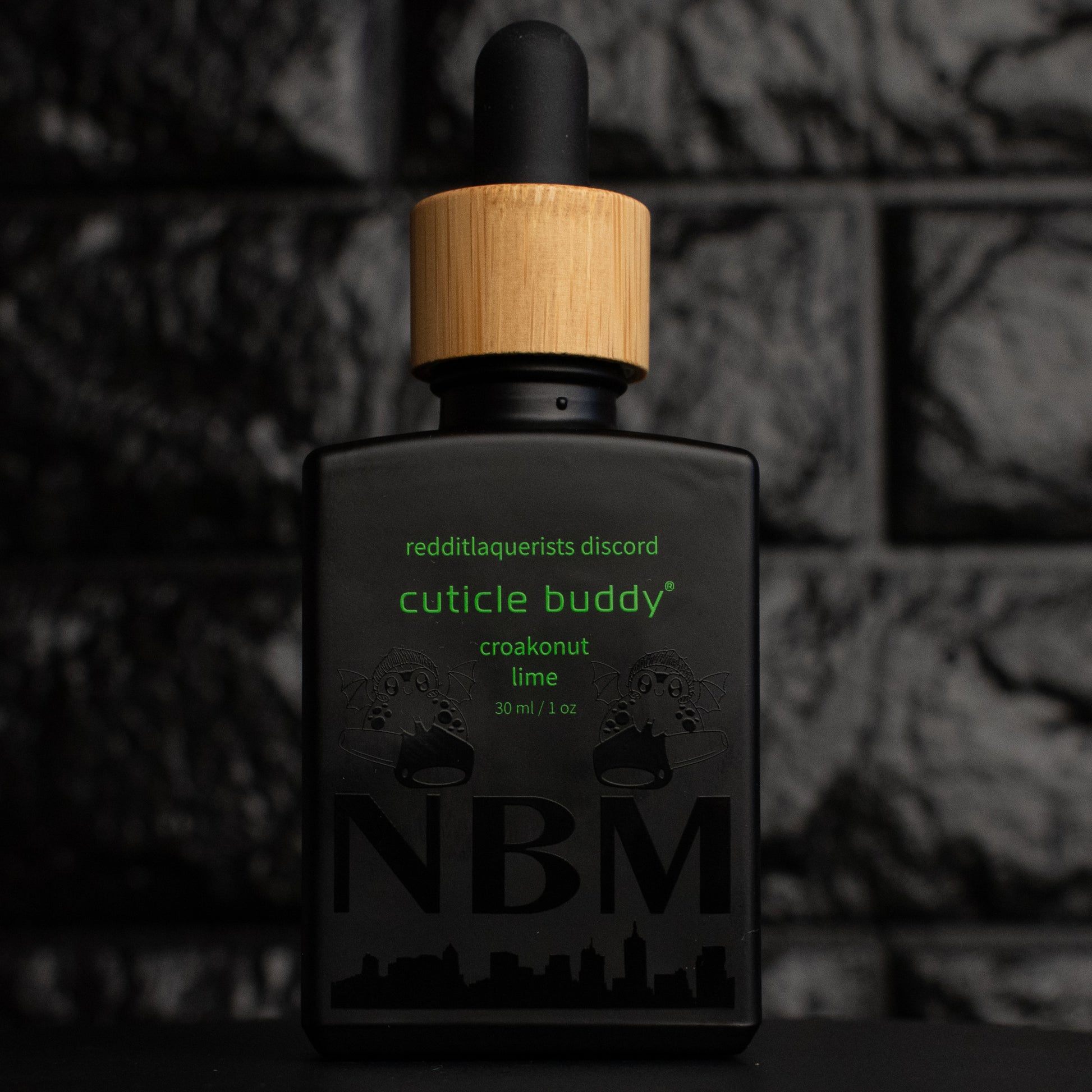 a 30ml matte black bottle of coconut lime verbena scented cuticle oil with a bamboo rubber dropper cap and little frog graphics on the bottle