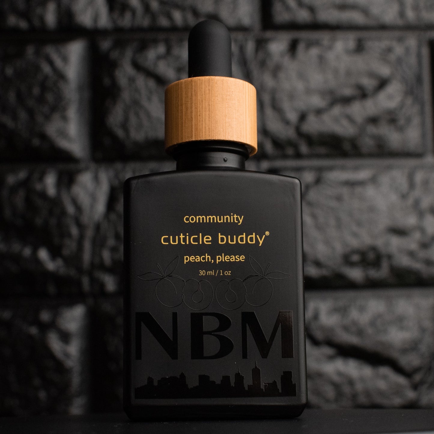 a matte black bottle marked community cuticle buddy peach, please 30ml/1oz with the NBM logo and an image of peaches printed on the bottle in black. there is a wooden rubber headed glass dropper in the bottle