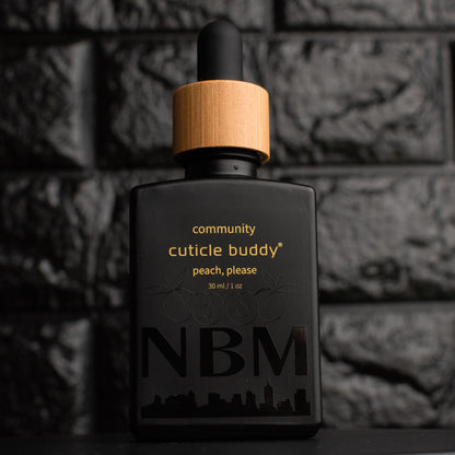 a matte black bottle marked community cuticle buddy peach, please 30ml/1oz with the NBM logo and an image of peaches printed on the bottle in black. there is a wooden rubber headed glass dropper in the bottle