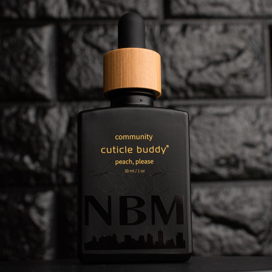a matte black bottle marked community cuticle buddy peach, please 30ml/1oz with the NBM logo and an image of peaches printed on the bottle in black. there is a wooden rubber headed glass dropper in the bottle