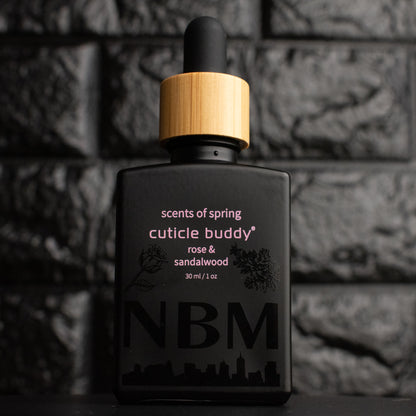a matte black bottle marked scents of spring cuticle buddy rose & sandalwood 30ml/1oz with the NBM logo and an image of a rose and sandalwood bush printed on the bottle in black. there is a wooden rubber headed glass dropper in the bottle