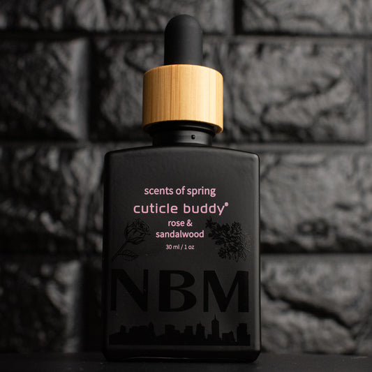 a matte black bottle marked scents of spring cuticle buddy rose & sandalwood 30ml/1oz with the NBM logo and an image of a rose and sandalwood bush printed on the bottle in black. there is a wooden rubber headed glass dropper in the bottle