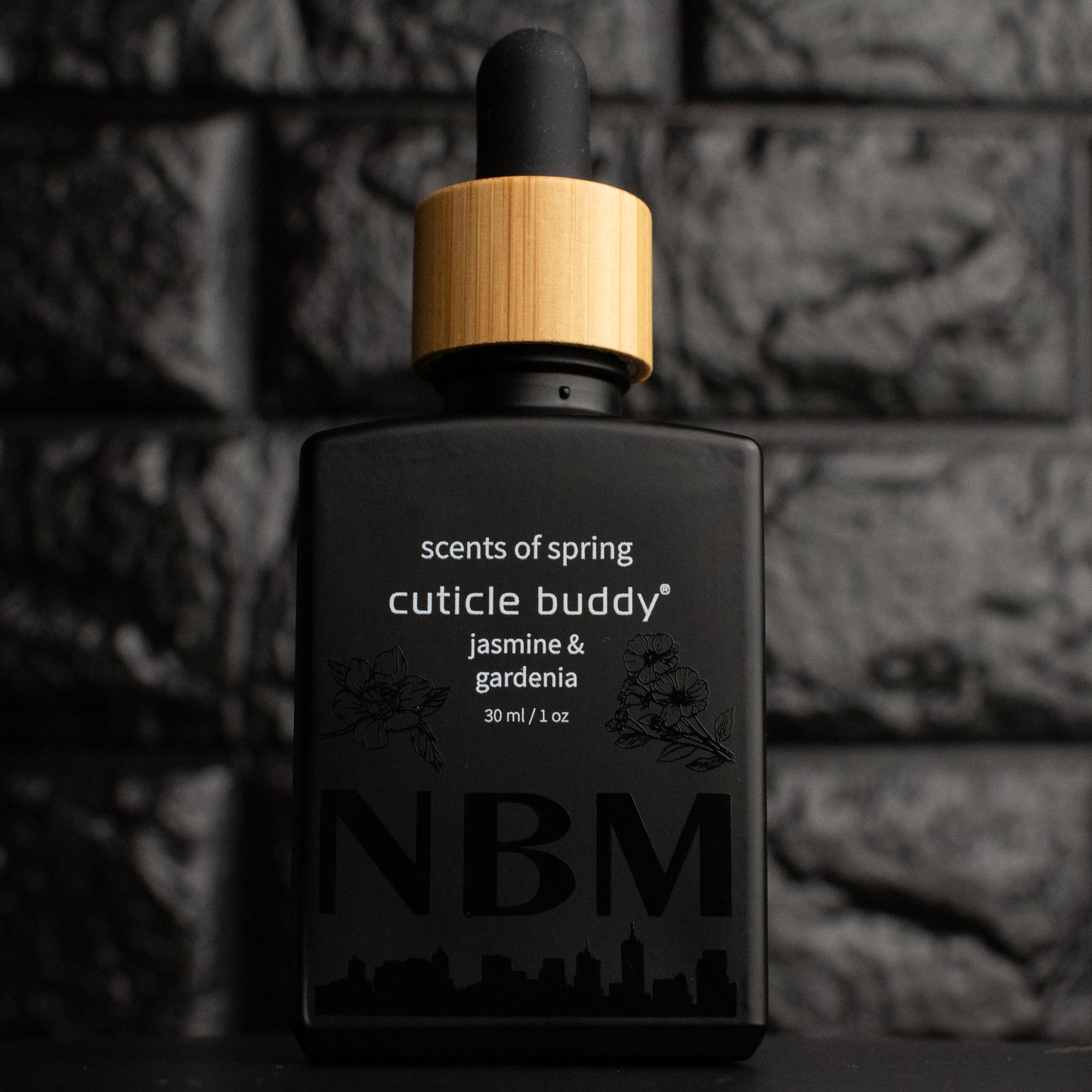 a matte black bottle marked scents of spring cuticle buddy jasmine & gardenia 30ml/1oz with the NBM logo and an image of a jasmine and gardenia printed on the bottle in black. there is a wooden rubber headed glass dropper in the bottle