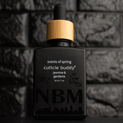 a matte black bottle marked scents of spring cuticle buddy jasmine & gardenia 30ml/1oz with the NBM logo and an image of a jasmine and gardenia printed on the bottle in black. there is a wooden rubber headed glass dropper in the bottle