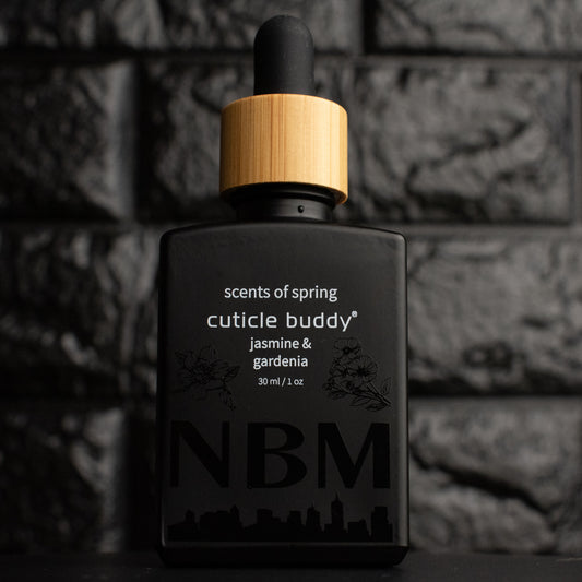a matte black bottle marked scents of spring cuticle buddy jasmine & gardenia 30ml/1oz with the NBM logo and an image of a jasmine and gardenia printed on the bottle in black. there is a wooden rubber headed glass dropper in the bottle