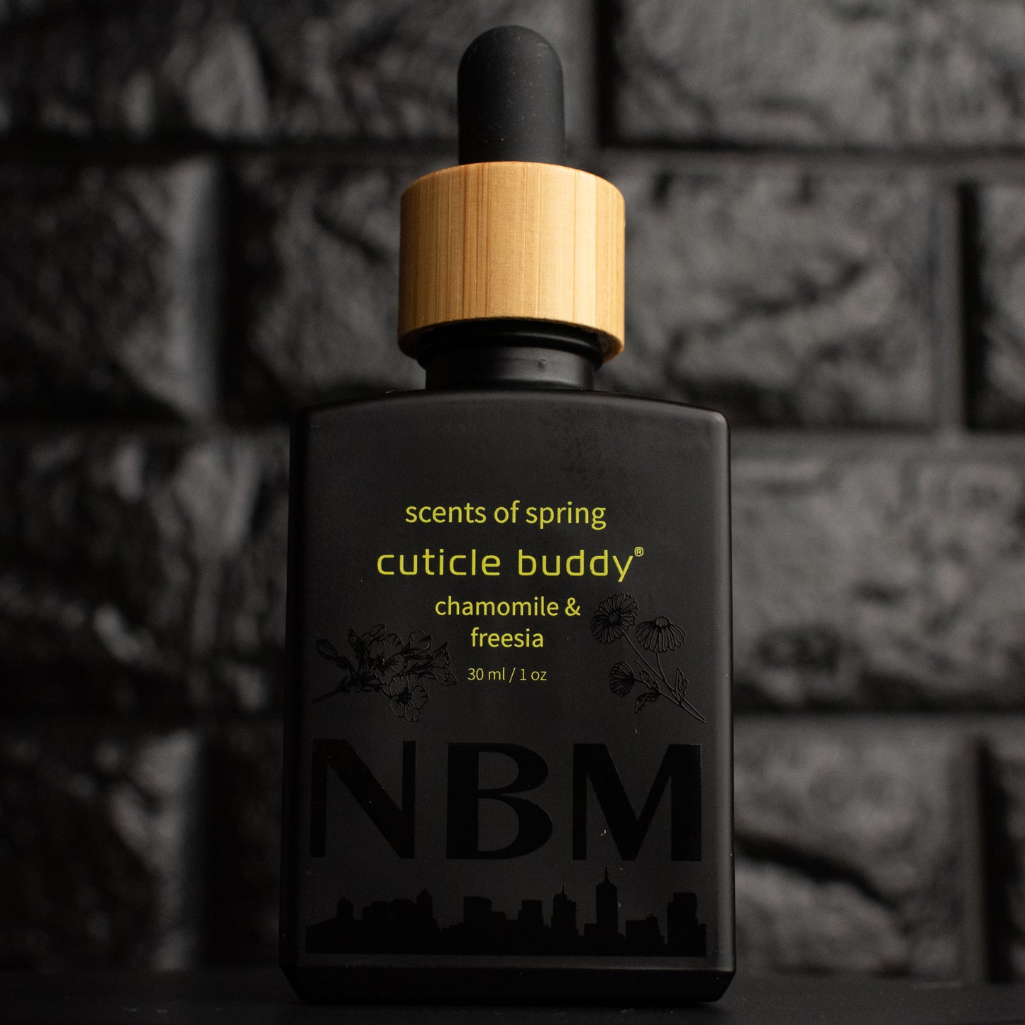 a matte black bottle of cuticle oil marked scents of spring cuticle buddy jasmine & gardenia 30ml/1oz with the NBM logo and an image of a chamomile and freesia flower printed on the bottle in black. there is a wooden rubber headed glass dropper in the bottle