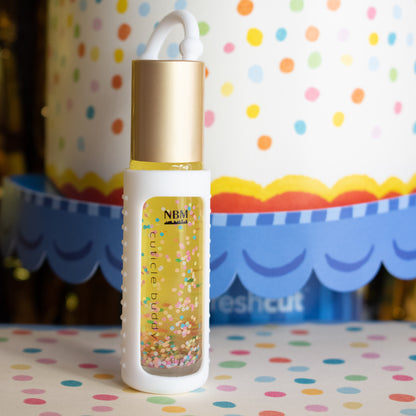 glass bottle of cuticle oil with a gold aluminum cap in a white silicone keychain holder and colorful biodegradable glitter floating inside in front of a paper birthday cake