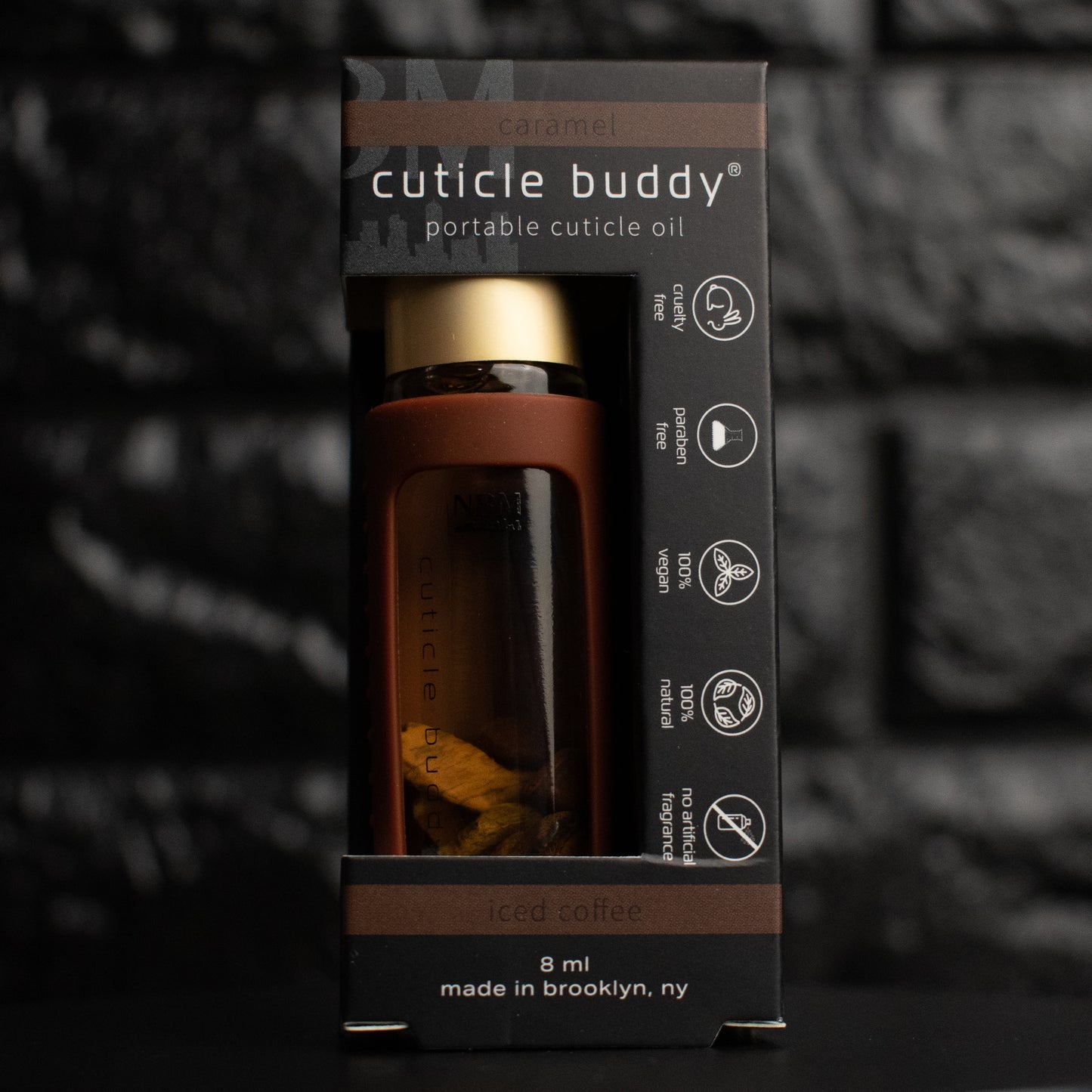 a glass bottle of oil with a gold aluminum cap in a black silicone holder on a black brick background. inside the bottle are black obsidian stones. in its packaging box