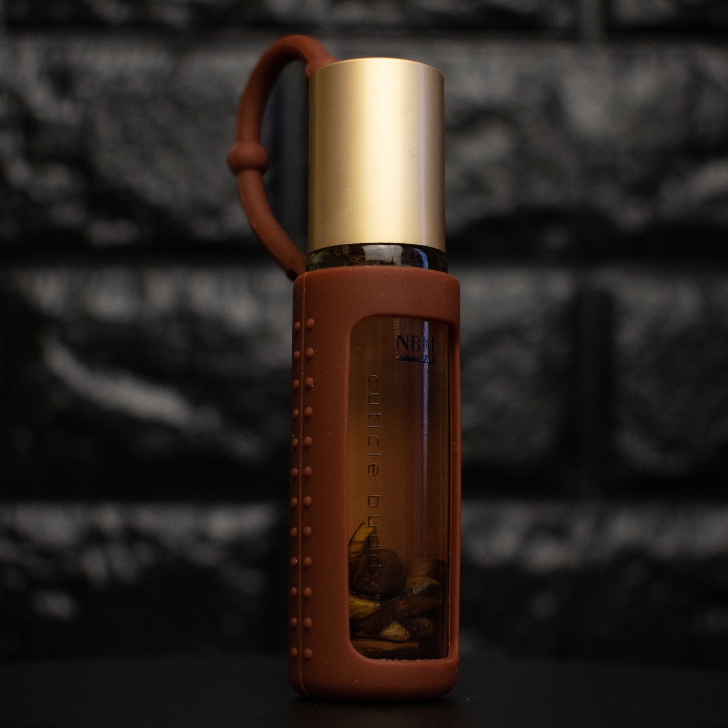 a glass bottle of oil with a gold aluminum cap in a brown silicone holder on a black brick background. inside the bottle are tigers eye stones