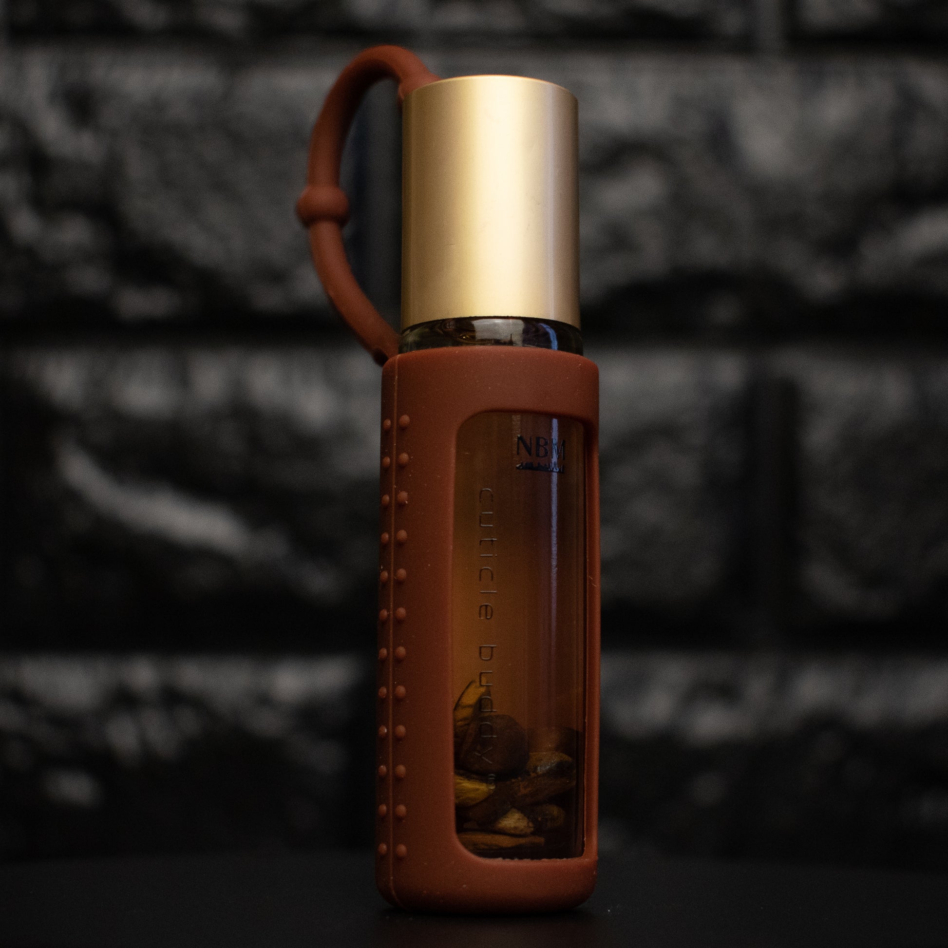a glass bottle of oil with a gold aluminum cap in a brown silicone holder on a black brick background. inside the bottle are tigers eye stones